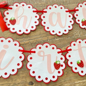 May include: A red and white banner with the word "Happy" spelled out in pink letters. Each letter is on a separate circle with a scalloped edge and red polka dots. There are small red strawberries with green leaves on each circle.