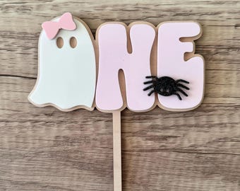 May include: Pink and white ghost with a pink bow, the word "ONE" in pink letters with a black spider on the "E", on a wooden stick. Halloween cake topper.