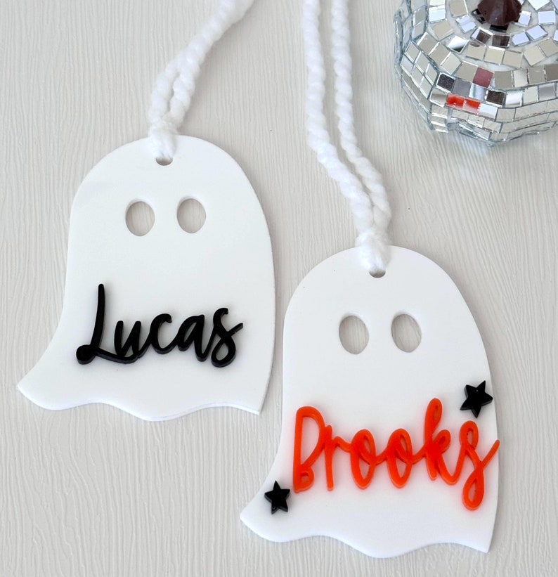 May include: Two white ghost-shaped ornaments with black and orange lettering. The first ghost has the name "Lucas" written in black. The second ghost has the name "Brooks" written in orange with two black stars.