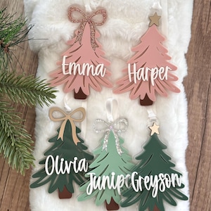 May include: Five personalised Christmas tree decorations with the names "Emma", "Harper", "Olivia", "Juniper", and "Greyson". The decorations are made of acrylic and have a pink and green colour scheme. Each decoration features a bow and a star.