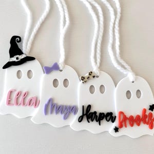 May include: Four white ghost-shaped ornaments with different coloured names and decorations. The names are Ella, Maya, Harper, and Brooks. One ghost has a witch's hat, one has a purple bow, one has a leopard print bow, and one has red stars.