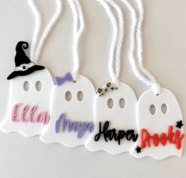 May include: Four white ghost-shaped ornaments with different coloured names and decorations. The names are Ella, Maya, Harper, and Brooks. One ghost has a witch's hat, one has a purple bow, one has a leopard print bow, and one has red stars.