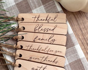 May include: Six wooden tags with twine attached. The tags are natural wood and have the following words engraved on them: thankful, blessed, family, thankful for you, give thanks, gather.