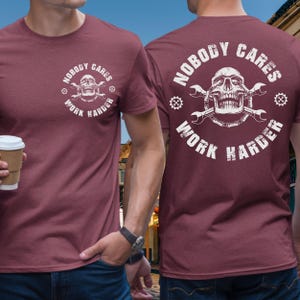 Puede incluir: Camiseta granate con un gráfico blanco en la parte delantera y trasera. El gráfico presenta una calavera, llaves inglesas y las palabras "NOBODY CARES WORK HARDER". La camiseta es de manga corta y tiene cuello redondo.