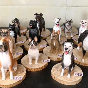 Op de afbeelding: Een verzameling hondenbeeldjes, met verschillende rassen zoals Boxers, Bulldogs en Huskies. Elk beeldje is met de hand beschilderd en staat op een klein houten schijfje. De collectie toont een verscheidenheid aan hondenrassen in verschillende poses.