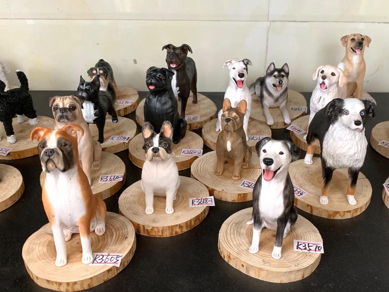Op de afbeelding: Een verzameling hondenbeeldjes, met verschillende rassen zoals Boxers, Bulldogs en Huskies. Elk beeldje is met de hand beschilderd en staat op een klein houten schijfje. De collectie toont een verscheidenheid aan hondenrassen in verschillende poses.