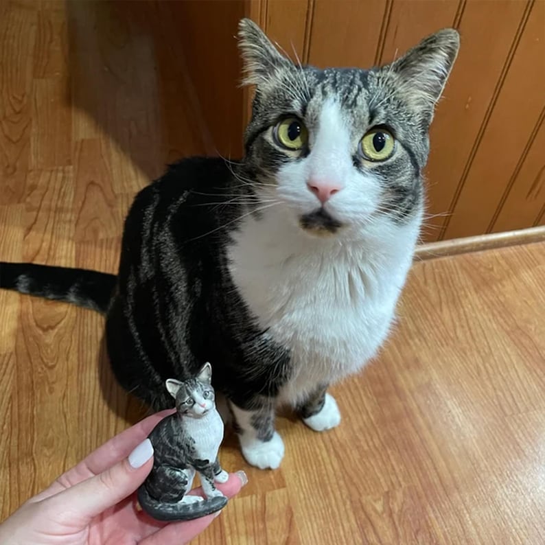 Op de afbeelding: Een tabby kat met witte aftekeningen zit naast een miniatuur keramieken kattenbeeldje. De kat heeft groene ogen en een roze neus. Het beeldje is een kleinere replica van de kat, met vergelijkbare markeringen.