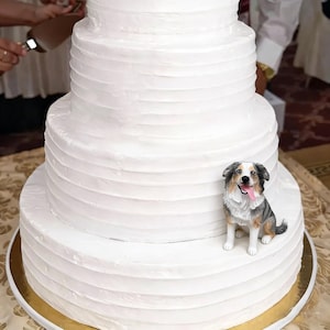 May include: A tiered white wedding cake with textured frosting. A small, detailed figurine of a dog with a pink tongue sits on the bottom tier. The cake is on a gold-colored cake stand.