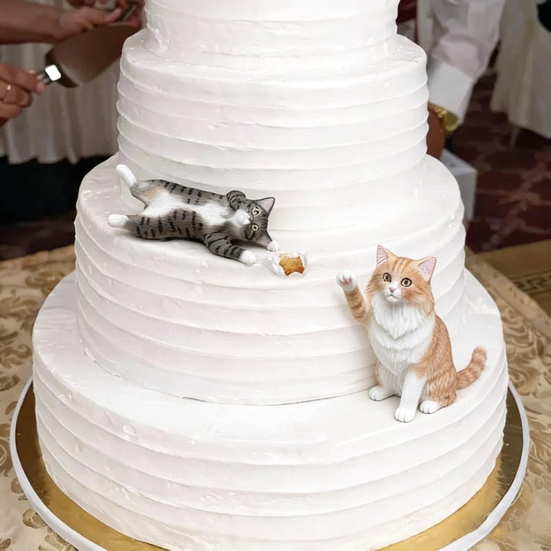 Op de afbeelding: Een witte bruidstaart met meerdere lagen, versierd met twee kattenbeeldjes. E&eacute;n kat is grijs en wit, de andere is oranje en wit. De taart heeft een botercr&egrave;me glazuur en staat op een gouden taartbord.