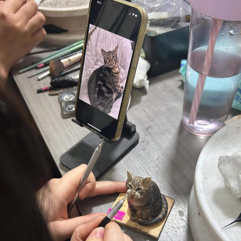 Op de afbeelding: Een gedetailleerde miniatuursculptuur van een tabbykat, bruin en grijs, staat op een kleine houten basis. De kunstenaar werkt aan de sculptuur, verwijzend naar een foto van een vergelijkbare kat op een smartphone. De sc&egrave;ne speelt zich af op een rommelig werkoppervlak.