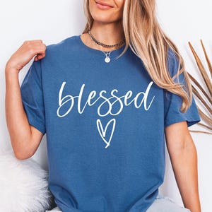 May include: Indigo blue t-shirt with the word "blessed" in white script and a white heart graphic. The shirt is a solid color and has short sleeves. The shirt is made of a soft material.