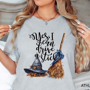 May include: An athletic heather light grey t-shirt featuring a graphic of a blue witch hat and a brown broomstick. The text reads, Yes, I can drive a stick! The design includes autumn leaves at the base of the hat and a blue fabric tie on the broom.