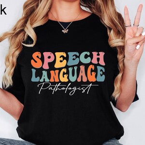 May include: Black t-shirt with the words "SPEECH LANGUAGE Pathologist" in colorful, retro-style lettering. The text is in orange, pink, blue, and yellow. The shirt is a crew neck and is being worn by a person.