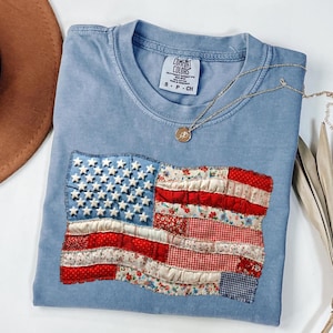 May include: A light blue Comfort Colors t-shirt with a sewn-on American flag design. The flag features white stars on a blue field and red and white stripes made of various patterned fabrics. A gold necklace rests on the shirt.
