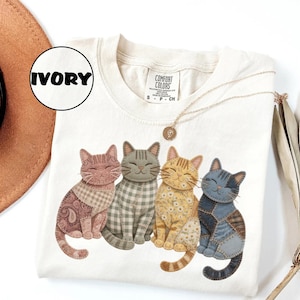 May include: An ivory short-sleeve t-shirt featuring a graphic of four sitting cats with patchwork-style patterns in shades of pink, gray, yellow, and blue. The shirt has a crew neckline and a Comfort Colors brand label at the inner neck area.