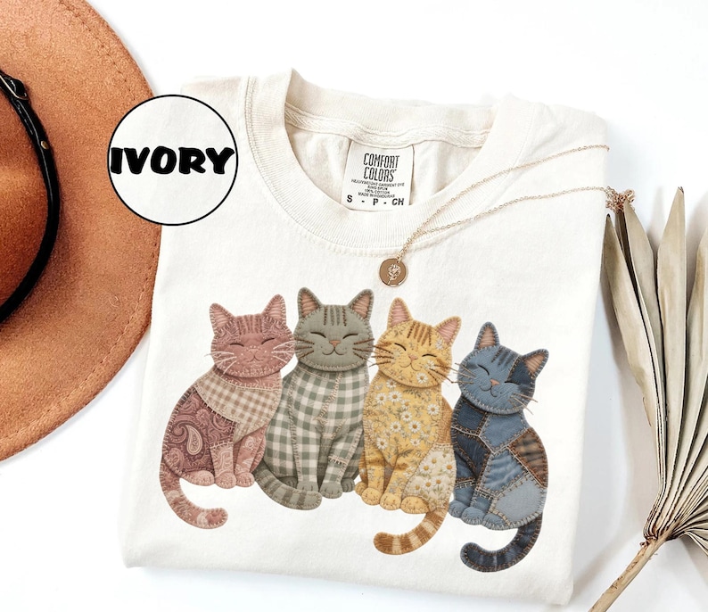 May include: An ivory short-sleeve t-shirt featuring a graphic of four sitting cats with patchwork-style patterns in shades of pink, gray, yellow, and blue. The shirt has a crew neckline and a Comfort Colors brand label at the inner neck area.