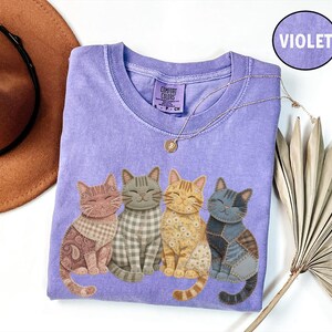 May include: A violet short-sleeve t-shirt featuring a graphic of four cats with different fabric-style patterns including paisley, gingham, floral, and patchwork. The shirt has a crew neckline and a Comfort Colors brand label at the inner neck.