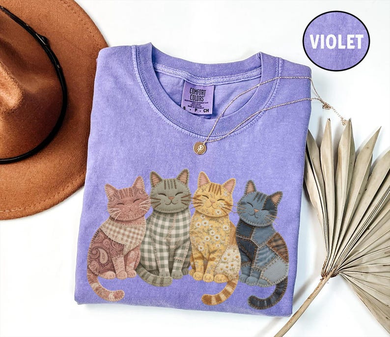 May include: A violet short-sleeve t-shirt featuring a graphic of four cats with different fabric-style patterns including paisley, gingham, floral, and patchwork. The shirt has a crew neckline and a Comfort Colors brand label at the inner neck.