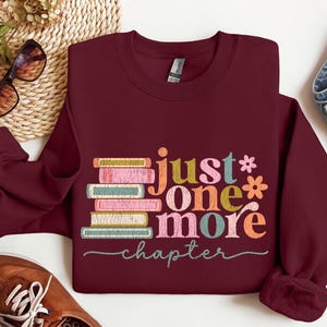 May include: A maroon sweatshirt with a stack of colorful books and the text "just one more chapter" printed on the front.