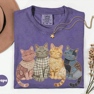 May include: A grape-colored t-shirt featuring a graphic of four cats with different patterned fur designs, including paisley, gingham, floral, and denim patchwork. The shirt has a Comfort Colors label at the neck and is size XL.