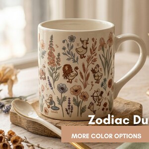 May include: Cream ceramic mug with a Zodiac Duck design, featuring floral and zodiac illustrations. The mug has a curved handle and is displayed with a spoon and a wooden coaster. Text on the mug reads "Zodiac Duck".