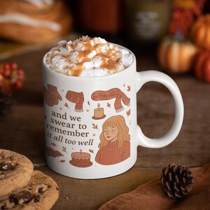 Könnte beinhalten: Weiße Keramiktasse gefüllt mit einem Getränk mit Schlagsahne, bestreut mit Zimt und Karamell. Die Tasse zeigt herbstliche Illustrationen und den Text "and we swear to remember it all too well". Schokoladen-Chip-Cookies und ein Tannenzapfen sind im Vordergrund.