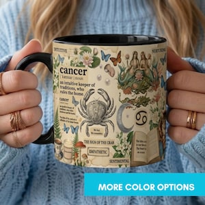 May include: A black-handled coffee mug featuring a Cancer zodiac design. The mug displays a crab illustration, mermaid figures, and botanical elements. Text includes "cancer" and "The Sign of the Crab". The mug is held by a person wearing a blue sweater.