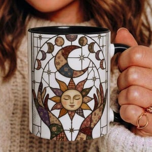May include: A white ceramic mug with a black interior and handle, featuring a stained-glass style graphic of a sun with a human face, a crescent moon, and stars, all rendered in a patchwork of muted earth tones and floral patterns.