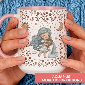 May include: A white ceramic mug with a pink handle and interior, featuring a cartoon illustration of a girl with long blue hair pouring water from a jug. The design includes small flowers, a sun, a moon, stars, and the Aquarius zodiac symbol.