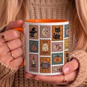 May include: A white ceramic mug with an orange handle and interior, featuring a grid of postage stamp-style illustrations including a witch, crescent moon, black cat, ghost with the text Cute Creepy, Capricorn symbol, candle, and pumpkin.
