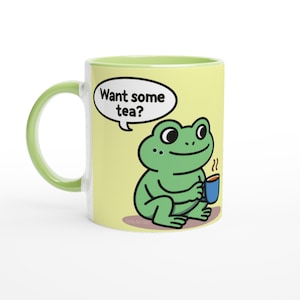 Puede incluir: Una taza de cerámica con asa e interior verdes. La taza presenta una rana de dibujos animados sosteniendo una taza de té azul, con el texto "Want some tea?" en un bocadillo. La taza tiene un fondo amarillo claro.