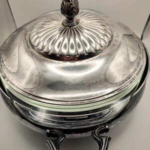 Könnte beinhalten: Eine silberfarbene Servierschale mit Glaseinsatz. Die Schale hat einen Deckel mit dekorativem Knauf und Griffen auf beiden Seiten. Der Deckel hat ein geriffeltes Design. Die Schale ist wahrscheinlich aus Metall und zum Servieren von Speisen bestimmt.