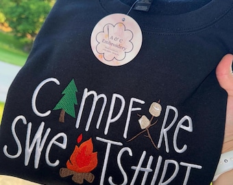 Puede incluir: Sudadera negra con texto bordado blanco y gráficos temáticos de acampada. El texto dice "Campfire Sweatshirt" con un árbol, una fogata y malvaviscos. Se adjunta una etiqueta redonda con el logotipo "A & C Embroidery".