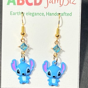 May include: A pair of gold-colored dangle earrings featuring blue cartoon character charms with purple ears. Each earring has a gold hook, a small gold bead, a square blue gem, and a blue charm. The earrings are displayed on a white card with green and red text.