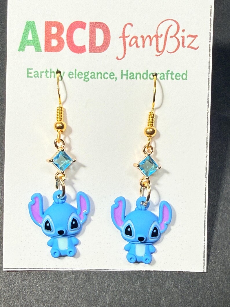 May include: A pair of gold-colored dangle earrings featuring blue cartoon character charms with purple ears. Each earring has a gold hook, a small gold bead, a square blue gem, and a blue charm. The earrings are displayed on a white card with green and red text.