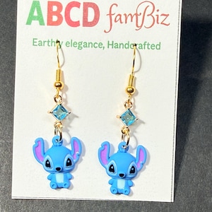May include: A pair of gold-colored dangle earrings featuring blue cartoon character charms. Each earring has a gold hook, a small gold bead, a square blue gem, and a blue character charm with purple ears. The earrings are displayed on a white card with text.