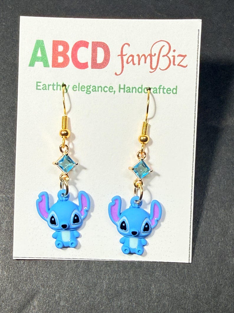 May include: A pair of gold-colored dangle earrings featuring blue cartoon character charms. Each earring has a gold hook, a small gold bead, a square blue gem, and a blue character charm with purple ears. The earrings are displayed on a white card with text.
