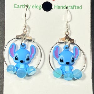 Puede incluir: Pendientes de aro plateados con dijes de Stitch azules, orejas rosas y detalles de cuentas azules. Los pendientes cuelgan de ganchos plateados y se exhiben en una tarjeta blanca con texto verde y rojo.