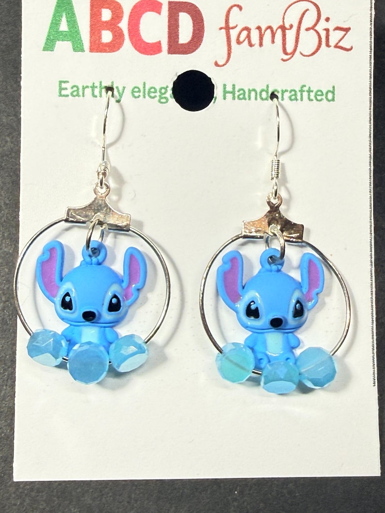 Puede incluir: Pendientes de aro plateados con dijes de Stitch azules, orejas rosas y detalles de cuentas azules. Los pendientes cuelgan de ganchos plateados y se exhiben en una tarjeta blanca con texto verde y rojo.