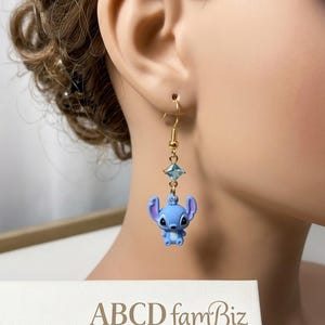 May include: A gold-colored earring with a blue cartoon character and a blue square gem. The earring is hanging from a person's ear. The text "ABCD famBiz" is visible in the lower part of the image.