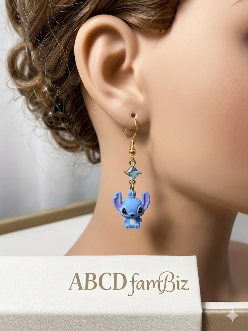May include: A gold-colored earring with a blue cartoon character and a blue square gem. The earring is hanging from a person's ear. The text "ABCD famBiz" is visible in the lower part of the image.