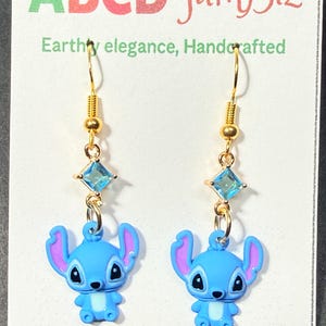 May include: A pair of gold-colored dangle earrings. Each earring features a blue cartoon character charm with purple ears, a light blue square gem, and a gold-colored hook. The earrings are displayed on a white card with the text "ABCD famBiz".