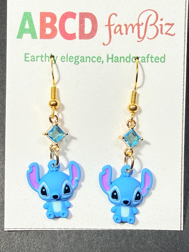 May include: A pair of gold-colored dangle earrings. Each earring features a blue cartoon character charm with purple ears, a light blue square gem, and a gold-colored hook. The earrings are displayed on a white card with the text "ABCD famBiz".