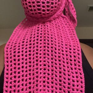 May include: A bright pink, open-weave crochet head covering and scarf. The head covering is rounded, and the scarf portion is rectangular. The crochet pattern features evenly spaced, square-shaped holes throughout the fabric.