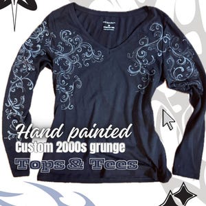 May include: A black long-sleeved V-neck top with hand-painted, pale blue, swirling floral patterns on the sleeves and chest. The top has the text "Hand painted Custom 2000s grunge Tops & Tees".