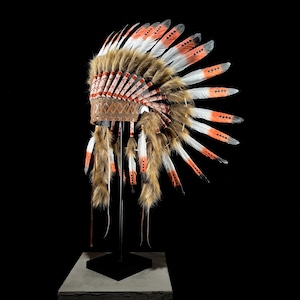 May include: A Native American headdress with brown fur, white and orange feathers, and a leather band. The headdress is displayed on a black stand against a black background. The feathers are tipped with orange and white accents.
