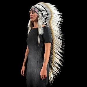 May include: A Native American headdress with white, brown, and black feathers. The headdress has a black and red beaded band. The feathers are arranged in a fan shape. The headdress is worn by a person wearing a black dress.