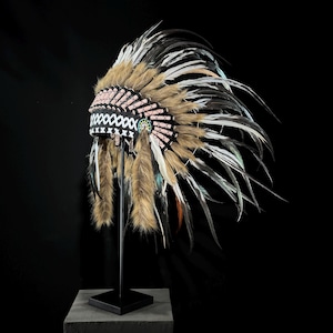 May include: A Native American headdress with a black and white beaded band, pink accents, and a multitude of feathers in shades of brown, white, and black. The headdress is displayed on a black stand against a black background.