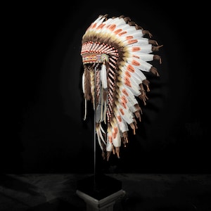 Op de afbeelding: Een traditionele Indiaanse hoofdtooi met witte, bruine en oranje veren. De hoofdtooi heeft gedetailleerd kralenwerk en wordt tentoongesteld op een zwarte standaard tegen een donkere achtergrond. De veren zijn in een waaier vorm gerangschikt.