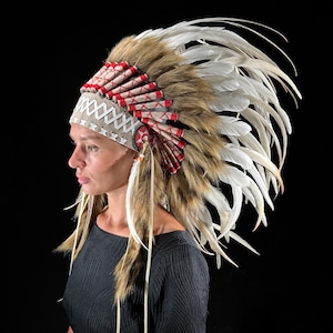 May include: A Native American headdress with a tan and red beaded headband, brown fur, and white feathers. The headdress is worn by a person against a black background. The feathers are long and flowing.